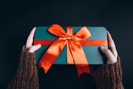 Woman's arms in brown sweater holding gift wrapped in green paper with orange bow on black backdrop