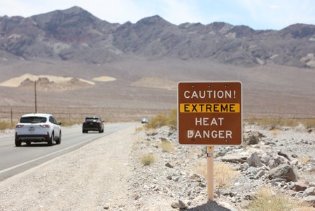 A heat advisory sign reading CAUTION EXTREMEHEAT DANGER next to a road with cars in Death Valley