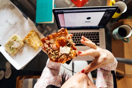 Overhead view of woman sitting in the kitchen at the glass table with laptop in headphones and holding pizza and coffee