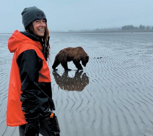 A woman looking at the camera, with a bear walking in the background